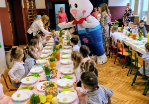 grupka przedszkolaków, nauczycielki, duży wielkanocny zając stół z wielkanocnymi potrawami