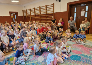 sala przedszkolna, duża grupa siedzących przedszkolaków, osoby dorosłe