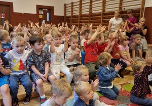 sala przedszkolna, duża grupa siedzących przedszkolaków, osoby dorosłe
