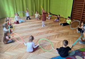 sala gimnastyczna, siedzące przedszkolaki z hula hop w rękach, dziewczynka w środku okręgu