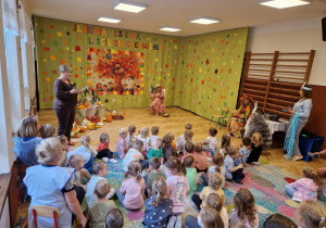 sala przedszkolna, jesienna dekoracja, nauczycielki w przebraniach do ról zająca, wiatru, lata, jesieni ,narratora, grupa siedzących na dywanie przedszkolaków