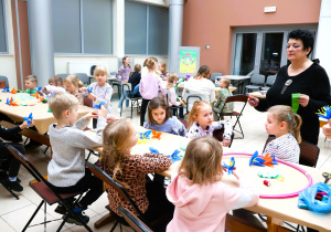 grupa dzieci, osoby dorosłe, przy stolikach, kwiaty z bibuły